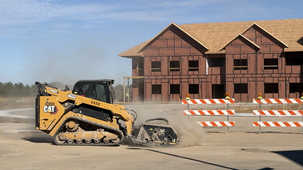 A yellow bulldozer uses a brush attachment to sand down poured concrete in front of a framed condo structure 