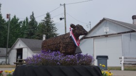 A small statue of a cow sits on top of a large tractor tire. Tiny purple and white flowers surround the statue, having been grown inside the tire. Yellow and red flowers can also be seen in separate pots surrounding the tire. Behind the mock up can be seen two white barns, one on the right of the statue and the other on the left. The left barn's door sits open while the one on the right is closed. The weather is overcast, as clouds fill the sky. 