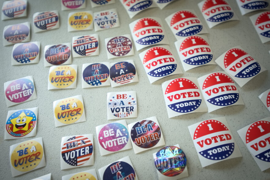 Close up image of about 30 different lapel stickers lying flat on a table. Some read "I Voted Today" with a red, white and blue design. Others read "Be a Voter" in colorful designs.