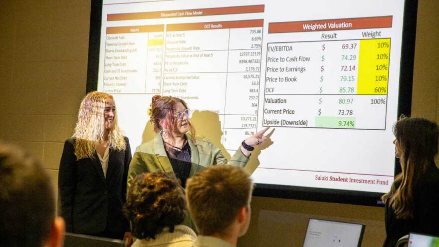 Three female students explain presentation at a projection screen in front of a classroom.