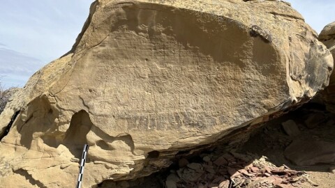 A measuring stick leans against a big boulder outside, which has Indigenous inscriptions on one of its sides.