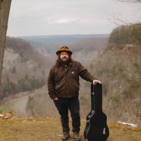 Tyler Westcott standing outside with his guitar case