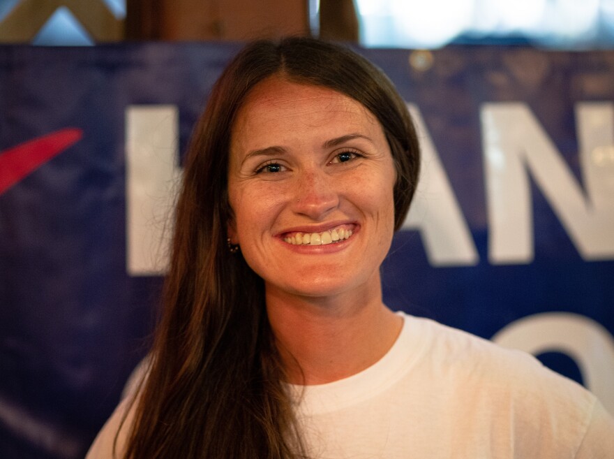 A woman with long brown hair smiles as she stands in front of a blue and white sign.