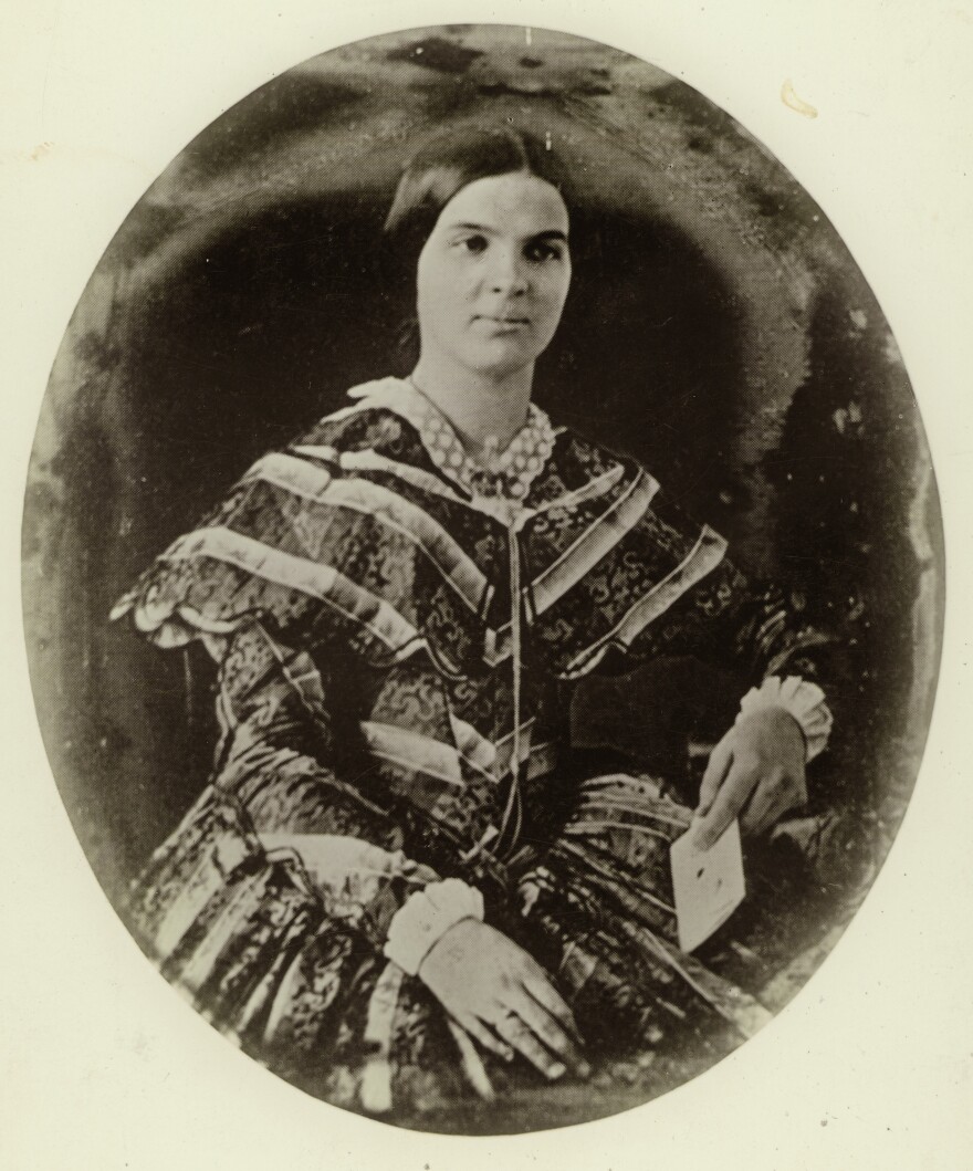 A photograph of a woman wearing a heavily patterned caplet-style dress with contrasting stripes. The collar of the dress is yet another pattern and forms a shallow V-neck. The cuffs appear to be made of lace. She sits in a chair, her left arm over the arm of the chair, holding a sealed envelope in her hand. Her right arm rests in her lap, and her wedding ring is visible on one of her long, elegant fingers. She has a calm face framed by dark hair parted in the middle and sleeked back. Her eyes are slightly downcast, her nose straight, and her full lips closed.