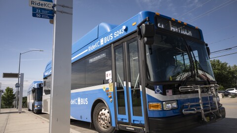 A blue bus appears to be moving away from a sidewalk. A sign near the bus reads "Ride KC, 21 Cleveland-Antioch; 50 Wornall-Brookside."