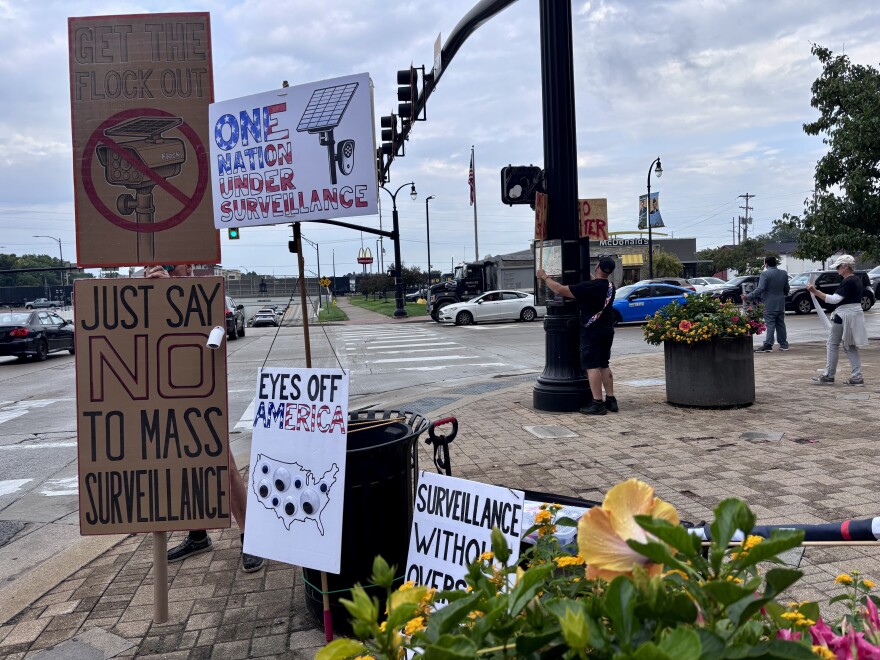 A man holds up several cardboard signs at an intersection as cars drive by. The signs have illustrations of surveillance cameras.