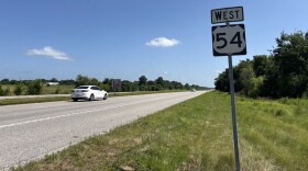 A white sedan drives down Highway 54. The photo is a wide shot of the road. A "West 54" road sign stands in the ditch. 