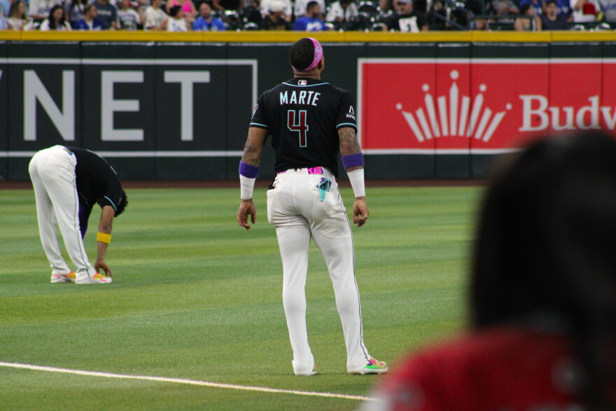 A baseball player stands on a field and looks up while a teammate stretches in the background