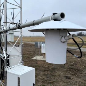 Weather monitoring gadgets are attached to a metal framework. The structure stands in the middle of a farm field.