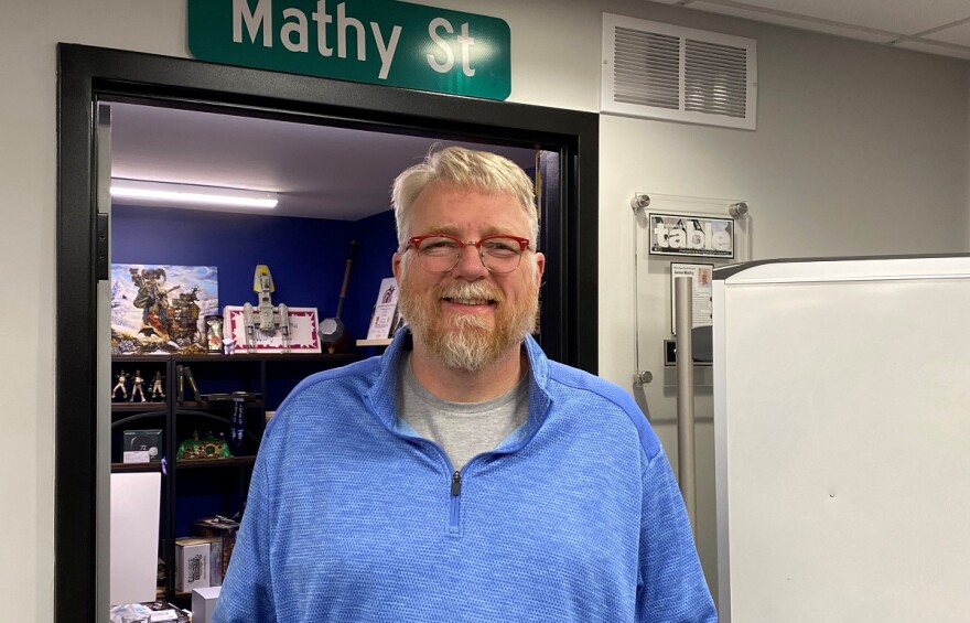 Man in blue quarter-zip standing in front of an office door with a street sign above that reads 'Mathy St.'
