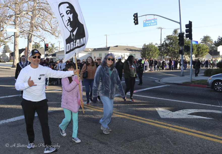 A large crowd marches in a city street on a sunny day. Two of the marchers, an adult and a child, are carrying a placard with the likeness of Martin Luther King, Jr. and the word 'Dream.'