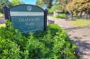 A sign for Trainsong park, along with bushes, sidewalk, picnic tables in the background.