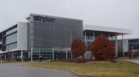 A tall building sits in front of a small smattering of short dark-orange-brown leafed trees. The building appears to be mostly glass, except for a see through metal mesh that climbs up the front of the facility, with "Stryker" sitting at the top of the mesh in black letters. Dark gray clouds cover the sky, denoting the rainy weather. 