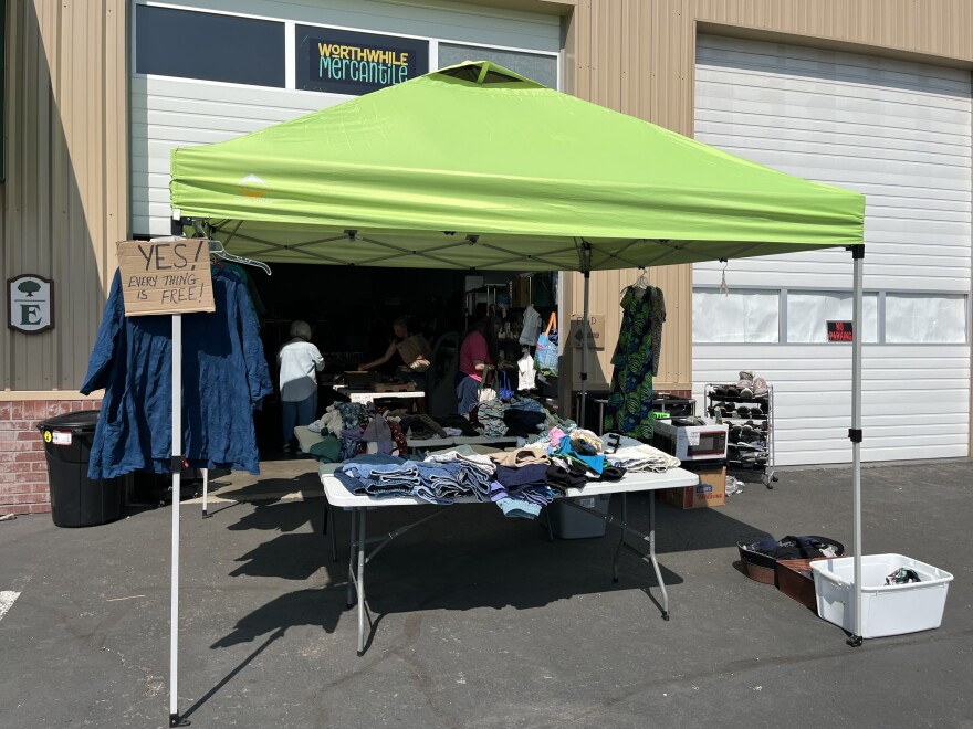A green tent canopy is set up in a parking lot, covering tables piled with clothes. 