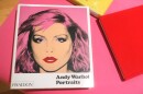 A glossy white book sits on a light-pink surface. The title reads: "Andy Warhol Portraits," and the cover shows a woman with red lipstick and pink hair. The corners of bright red and yellow books are also visible. 