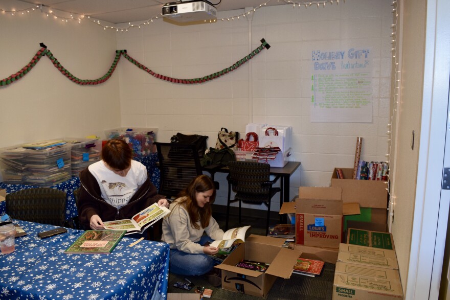 Two people look at picture books, in a room lined with boxes and plastic bins of books. One table is filled with empty gift bags.