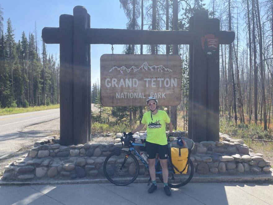Friederike Benning stands smiling with her bike in front of the Grand Teton National Park sign
