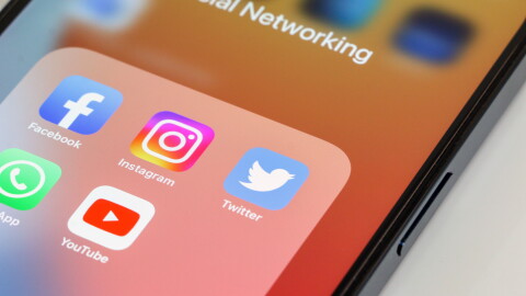 Close-up of popular social media app icons, Facebook, Instagram, Twitter, WhatsApp and YouTube, in that order, displayed on smartphone screen with orange wallpaper. Device is on white background.