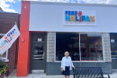A woman wearing a blue skirt and white cardigan stands in front of a building painted in bright orange on the side and the words "Feed Halifax" on the face of the building. 