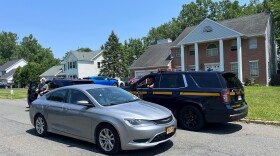 This photo is taken from a suburban street. Several houses, including the one Harbe disappeared from, are visible in the background. Two State Police vehicles are parked in front of the lawn on the street. A silver sedan is driving through the photo. 