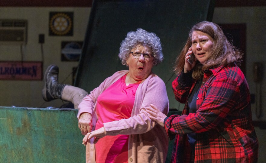 Two women on stage with a leg sticking out of a dumpster behind them.