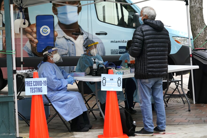 Healthcare workers sit at a white foldout table underneath a white tent. The workers wear medical gowns, face shields, and face masks. On the table are various medical supplies such as hand sanitizer and face masks. A man in jeans, a puffy jacket, and a surgical mask stands in front of the table. Two orange traffic cones are set up in front of the tent. A sign on one cone reads, "Free COVID-19 Testing." The other cone has a sign that reads, "Six feet. Keep your distance." Behind the healthcare workers is a large vehicle with the words, "Miami-Dade County," printed on it. 