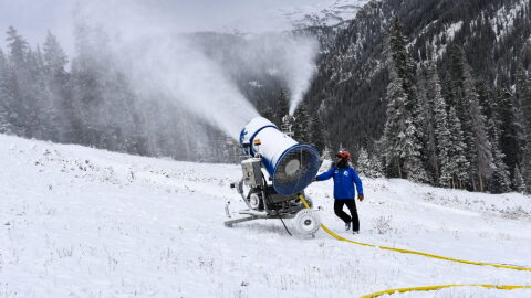 A large snowgun shoots snow on the side of a hill with pine trees in the background.