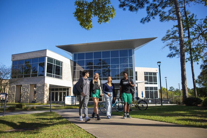 Four diverse students walk on a pathway with a UWF building and bicycles in the background