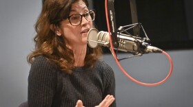 A woman sitting inside a radio studio talks at a microphone and gestures with her right hand.
