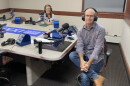 Two people wearing headphones and eyeglasses sit at a table in a radio talk studio: a woman at left has shoulder-length brown hair and is wearing a blue-and-white striped sweater with a blue collar; a balding man at right is wearing a plaid button-down shirt, blue jeans and brown shoes.