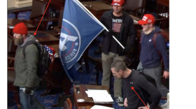 Joshua Hughes, wearing a red beanie, walks through an aisle in the Senate Chamber. Jerod Hughes stands nearby looking at papers atop a desk.