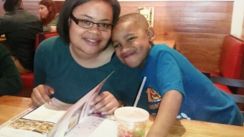 Atatiana Jefferson, a Black woman wearing black glasses and a teal shirt, smiles for a photograph with a young boy who makes a silly face. She's sitting in a restaurant and flipping through a menu.