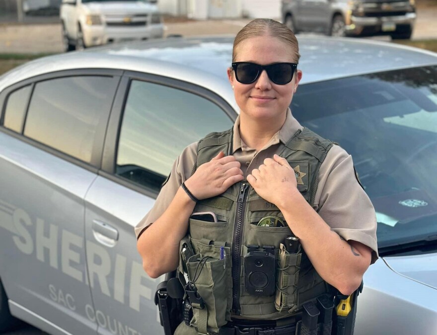 A woman is wearing a law enforcement uniform, sunglasses and is standing in front of a sheriff's car.