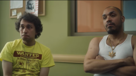 Alex sits in a yellow tee shirt with a red design and Dylan sits in a white cut off shirt and flashy gold chain.