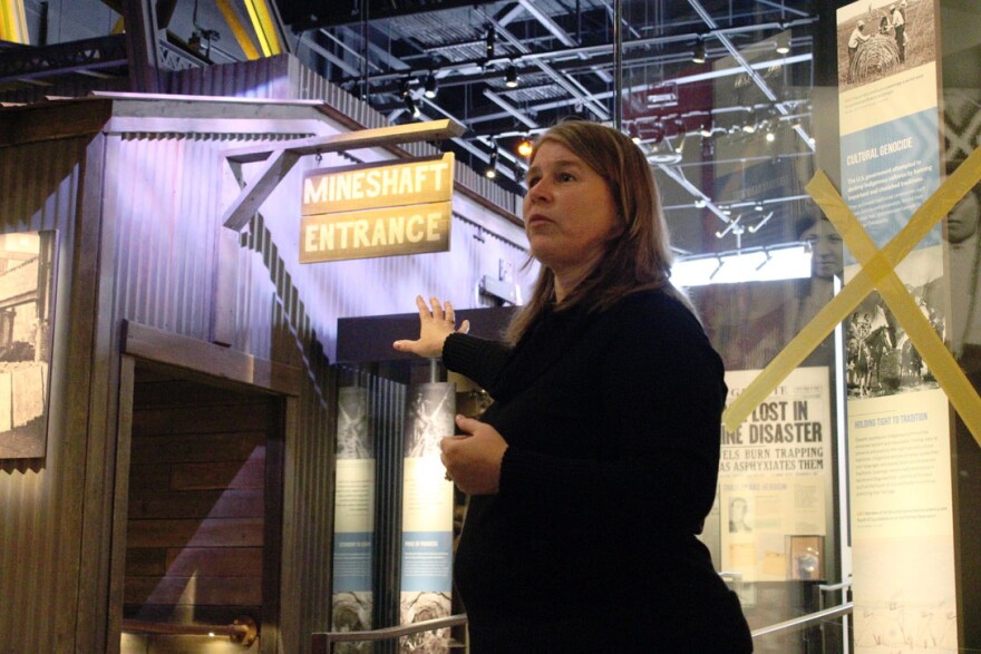 Molly Kruckenberg, executive director of the Montana Historical Society, gestures while leading a tour through the Montana Heritage Center. She stands near a life-size exhibit entrance labeled “Mineshaft Entrance,” with interpretive panels and historical images visible in the background.