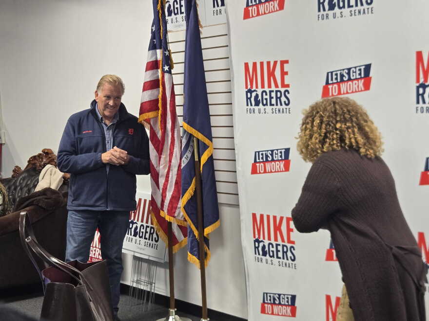A man in a navy blue zip-up jacket and jeans stands next to an American flag and a Michigan state flag, speaking to a woman with curly blonde hair who has her back to the camera.