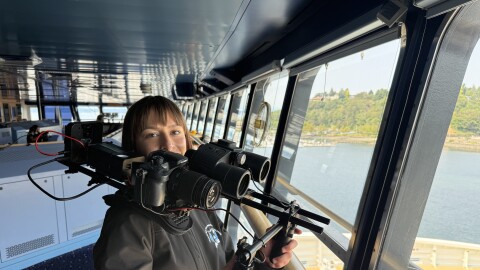 A woman wearing a black jacket standing next to the picture windows on the bridge of the MSC Poesia holds a large rig that combines binoculars and a video camera. 