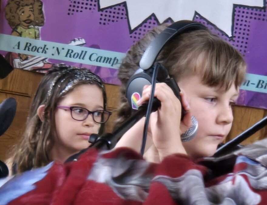 a close-up of two kids looking to their left. The child in the front has hands on a microphone. In the background, a poster says "A Rock N Roll Camp"