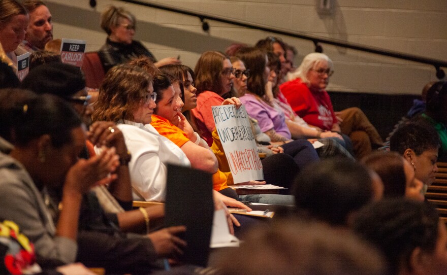 An attendee holds up a sign: "pre determined, underplanned, unacceptable."