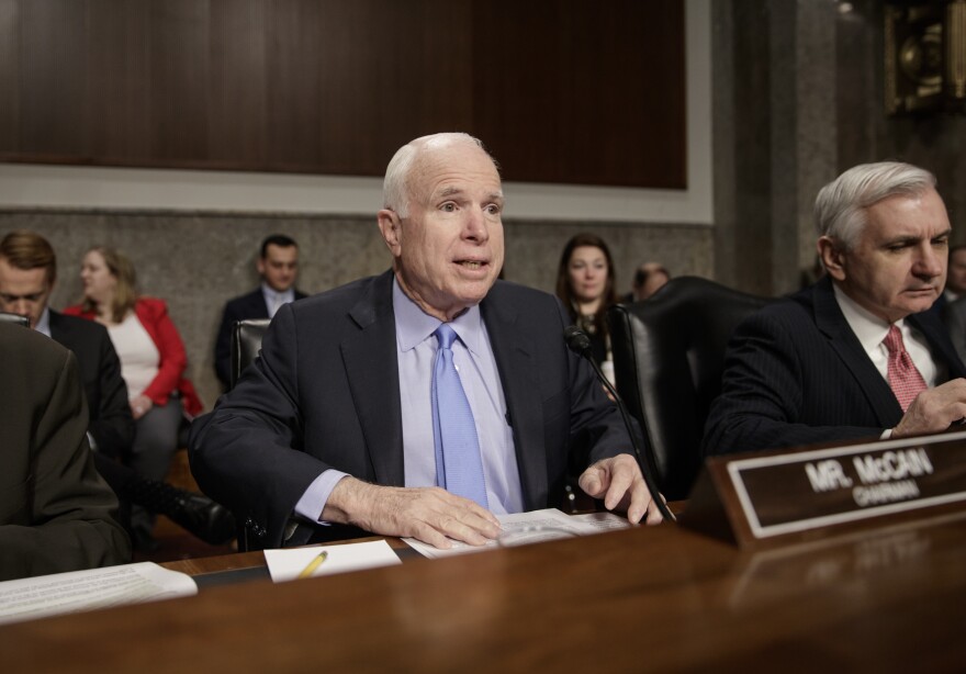 A man in a suit speaks during a Senate hearing while seated at a long desk with microphones and other officials around him. A nameplate reading “Mr. McCain” is visible in the foreground.