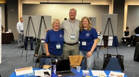 Three people stand in front of a table at a conference. The table reads Deer River Full Service Schools.