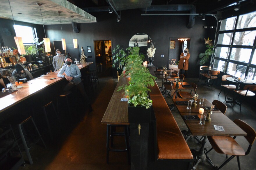 Wide shot of the interior of a restaurant. One person is leaning on the bar while sitting on a stool. Others are working behind the bar. A woman stands near the entrance. You can see a large glass rollup door on the right, six tables with chairs and a long row of ferns in the middle of the room.