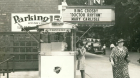 Image of the Irvin Theater in Bloomington. A woman buys popcorn from the stand outside. Another older woman in a long dress and hat walks by. The marquee sign is for Bing Crosby "Dr Rhythm" and Mary Carlisle.