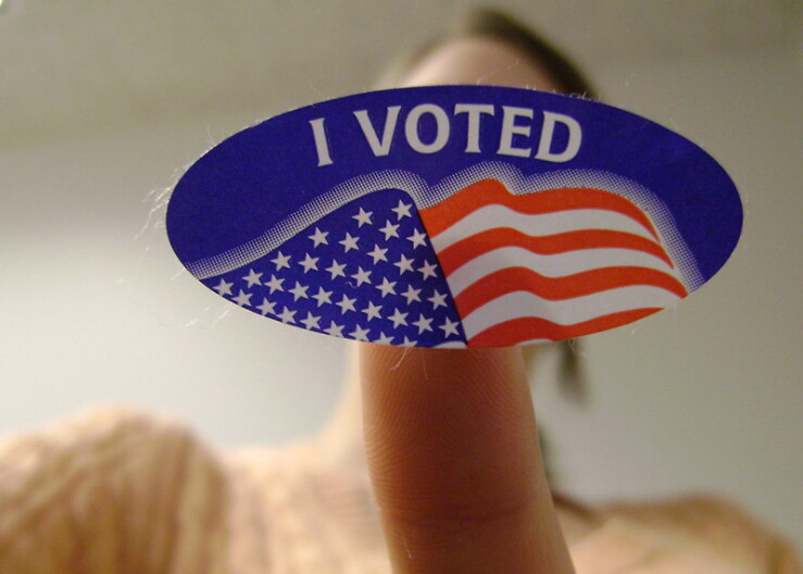 A finger holds up a blue "I voted" sticker with an American flag on it.