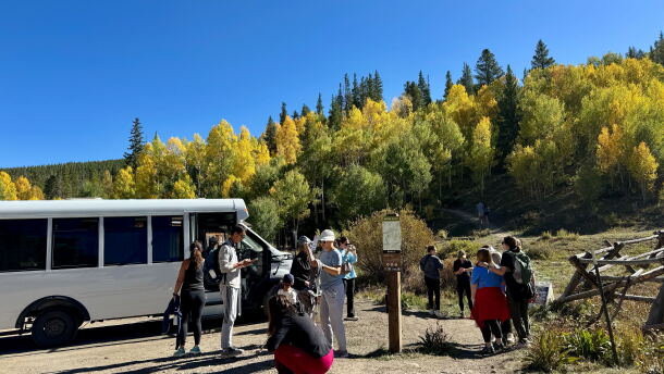 A shuttle bus drops off hikers in a dirt parking lot in front of a trail with yellow leaves