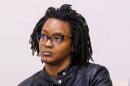 Black woman with glasses, nose piercing and short dreads wearing a black leather jacket and looking off in the distance