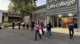 A group of people, including a man holding a tray of margaritas, walk out of the doors of Cafe College as mariachi play in the background next to the I-35 overpass.