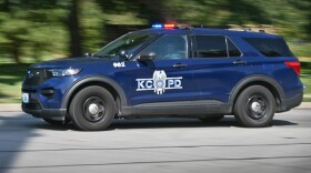 A blue police SUV with the "KCPD" logo moves on a city street with its emergency lights on during daylight hours. The background is blurred with motion.