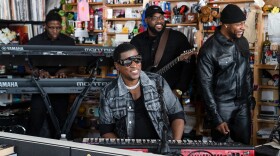 A man wearing sunglasses and a dark denim jacket smiles while seated behind a keyboard on an office desk with three musicians behind him, along with shelves full of music paraphernalia.