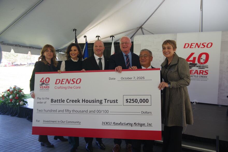 Three women and three men all hold up a large white check with red trim on the top and bottom. The amount is $250,000, paid to the Battle Creek Housing Trust, as written on the check. At the top right can be seen "DENSO," the company giving out the check, written in red letters. Below this reads their motto, "Crafting the Core." To the right of DENSO can be seen the words "40 years." The people holding the check are standing in front of a temporary stage, the back of which also has DENSO's name written on it along with the 40 year anniversary.